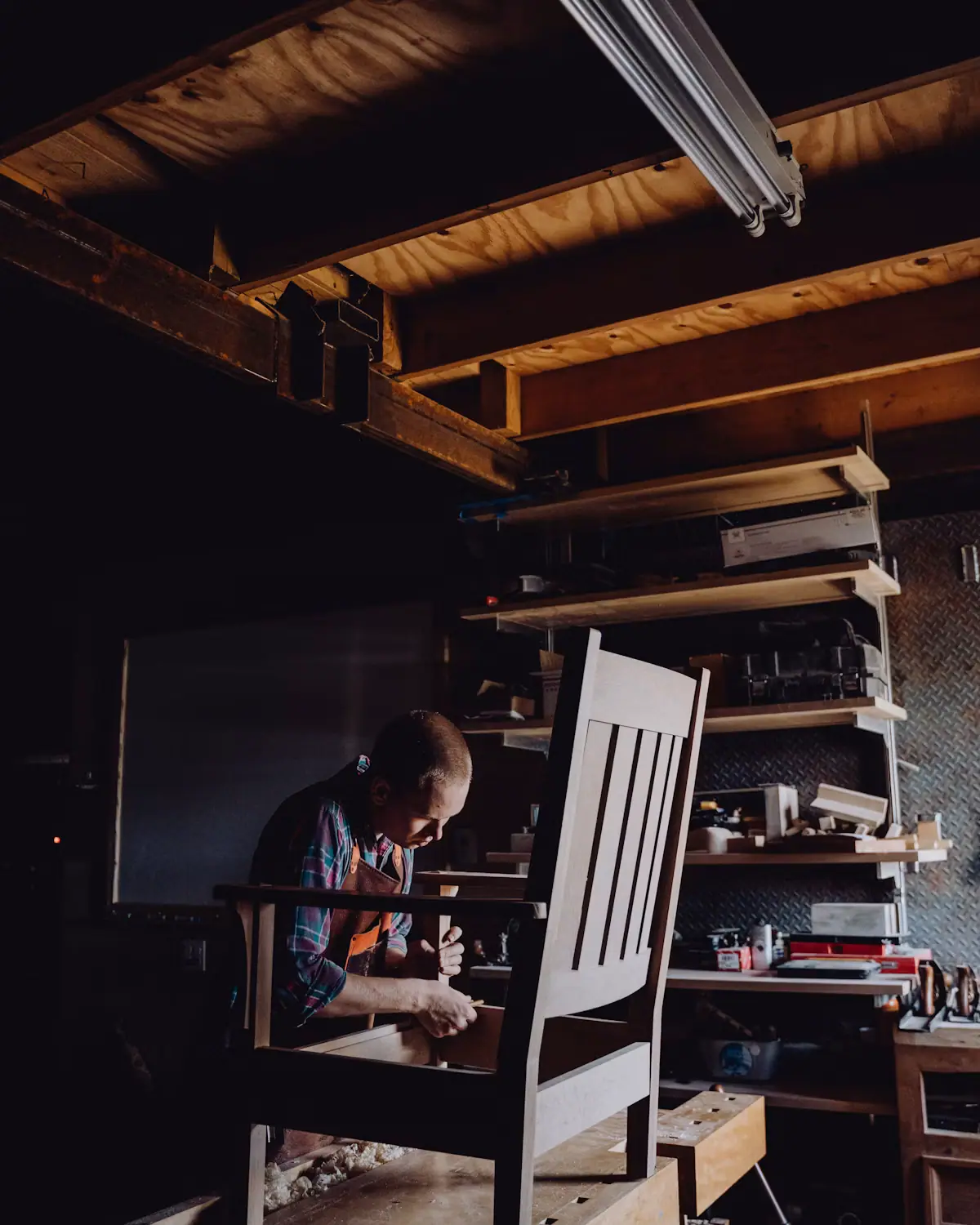 A maker working on a wooden chair frame at a bench in a timber-framed workshop under warm overhead light